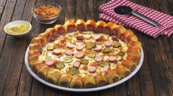 Eine Hotdog-Pizza auf einem Teller, daneben eine Schale mit Röstzwiebeln, eine mit Senf und ein Pizzaschneider auf einem Tuch.