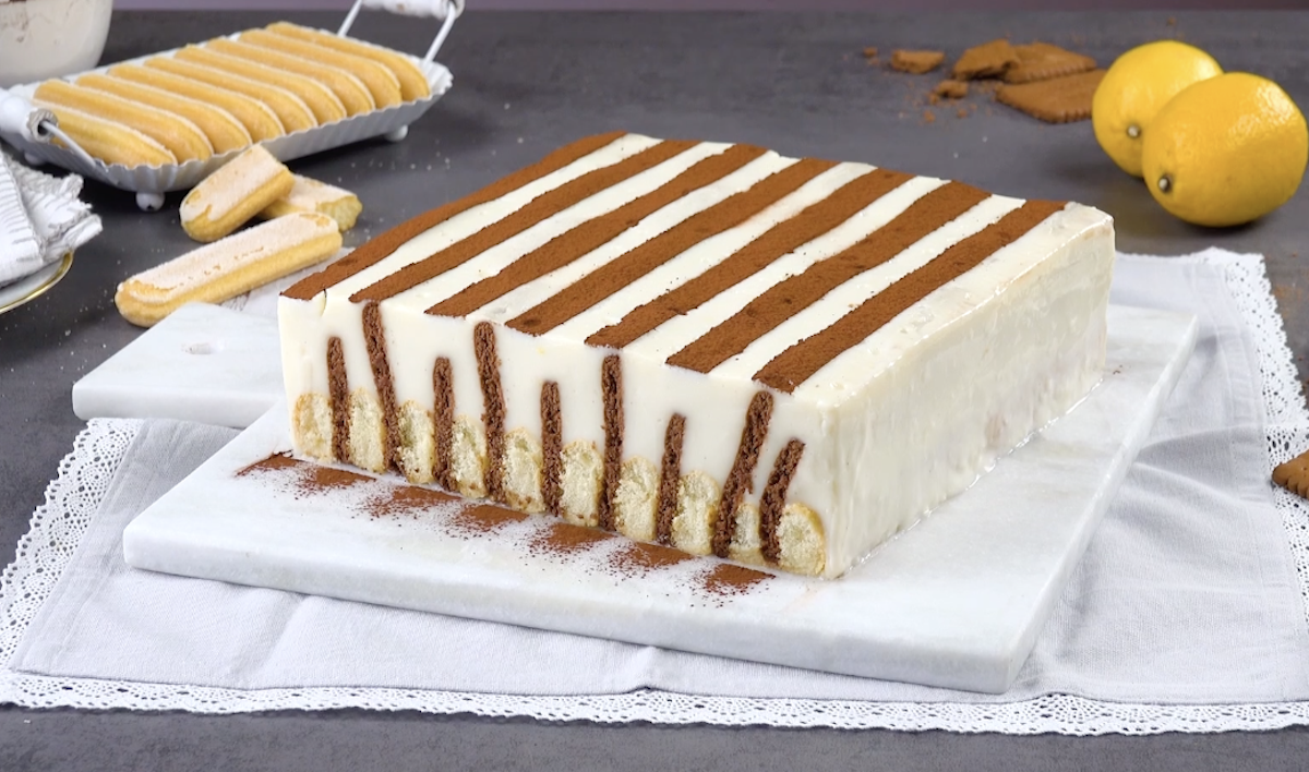 Ein Kekskuchen mit Pudding im Streifenlook auf einem Marmorbrett. Daneben zwei Zitronen und ein Tablett mit Löffelbiskuit.