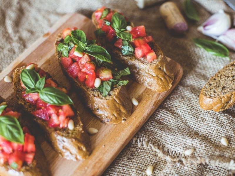 4 kleine Brote mit Bruschetta Italiana auf einem Holzbrett. Daneben liegen Brotscheiben.