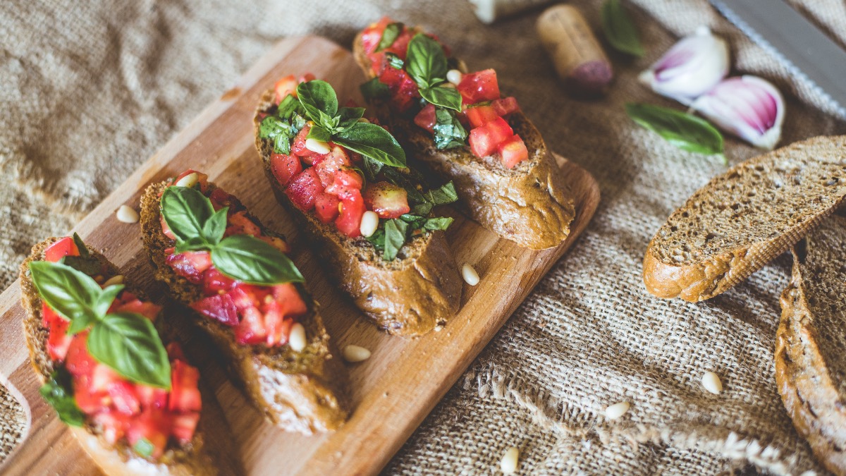 4 kleine Brote mit Bruschetta Italiana auf einem Holzbrett. Daneben liegen Brotscheiben.