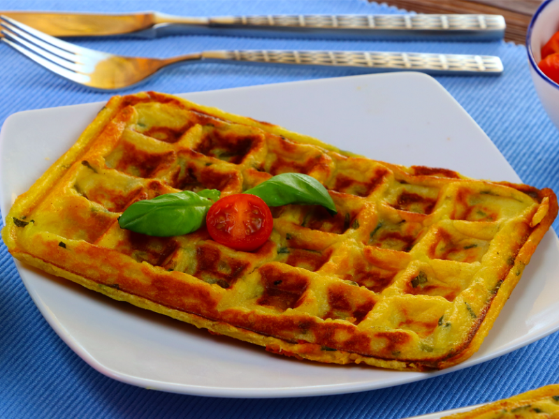 Eine gefüllte Kartoffelwaffel auf einem viereckigen Teller. Daneben steht eine Tasse Kaffee und eine Schale mit Mini-Tomaten. Im Hintergrund liegt Besteck.