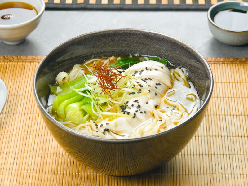 Ramen in einer Schüssel. Daneben stehen Schalen mit Soße und Besteck.