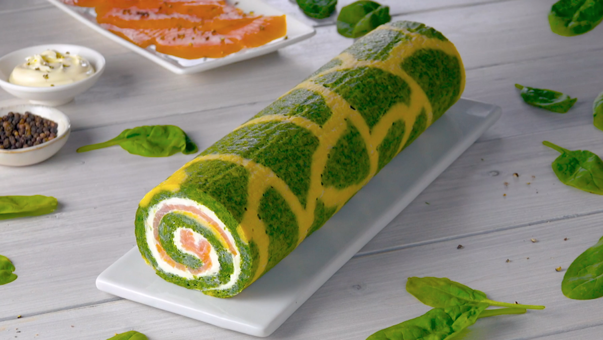 Eine Spinat-Lachs-Rolle auf einer Servierplatte. Drumherum wurden Spinatblätter und Schälchen mit Zutaten verteilt.