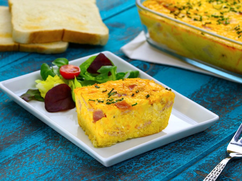 Eine Portion Toastbrot-Quiche mit Salat auf einem eckigen Teller. Im Hintergrund ist die restliche Quiche in der Auflaufform zu sehen.