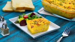 Eine Portion Toastbrot-Quiche mit Salat auf einem eckigen Teller. Im Hintergrund ist die restliche Quiche in der Auflaufform zu sehen.