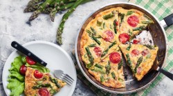 Eine spanische Tortilla mit grünem Spargel und Tomaten in der Draufsicht. Daneben steht ein Teller mit einem Stück Tortilla.