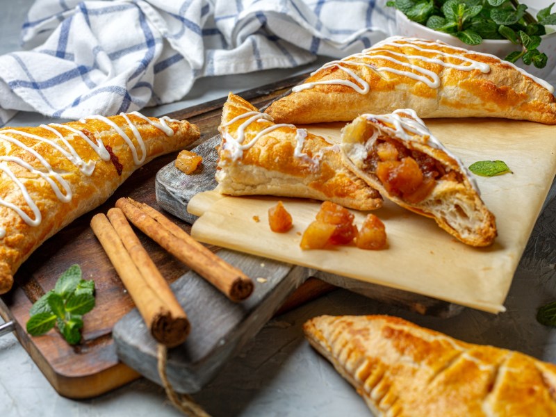 Ein paar Apfel-Kürbis-Taschen auf einem Brett, einige davon angebrochen, daneben Zimtstangen und ein Geschirrtuch.