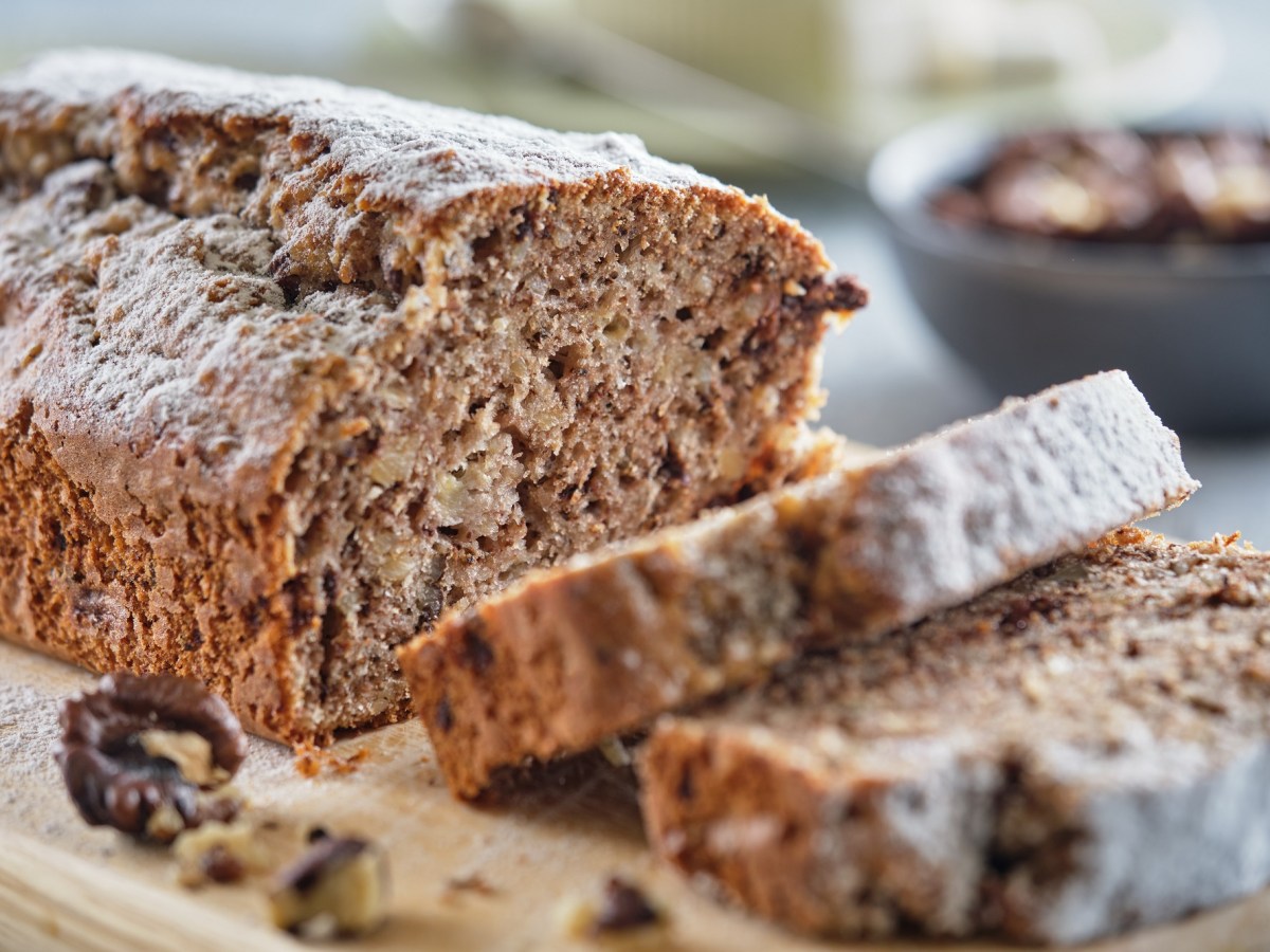 Ein angeschnittenes einfaches Dinkel-Vollkornbrot auf einem Brett, auf dem auch Walnüsse liegen, im Hintergrund unscharf Schüsseln mit Nüssen.