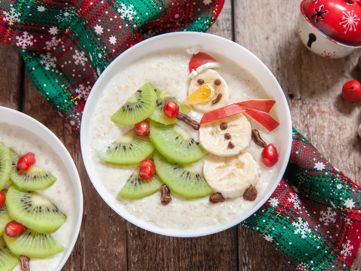 Zwei Schüsseln Weihnachtsmilchreis dekoriert mit Schneemännern und Weihnachtsbäumen aus Bananen und Kiwi, drumherum weihnachtliche Deko, Draufsicht.