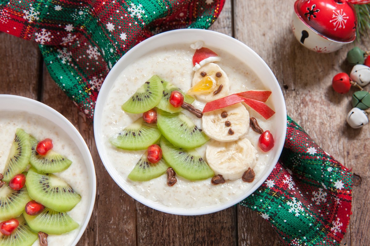 Zwei SchÃ¼sseln Weihnachtsmilchreis dekoriert mit SchneemÃ¤nnern und WeihnachtsbÃ¤umen aus Bananen und Kiwi, drumherum weihnachtliche Deko, Draufsicht.