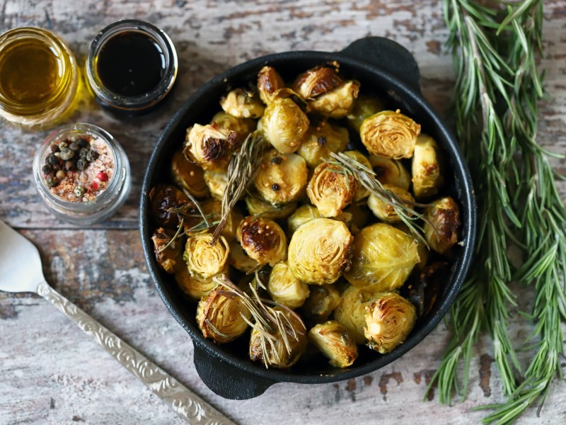 Gebackener Rosenkohl mit Balsamico und Ahornsirup in einer gusseisernen Form in der Draufsicht, daneben Gläser mit Zutaten und Rosmarin.