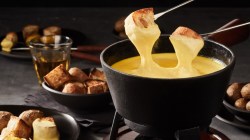 Ein Käsefondue, in das zwei Brotstücke getaucht werden. Im Hintergrund stehen Schalen und Teller mit Brot und ein Glas mit Wein.