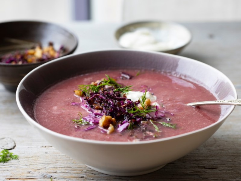 Ein Teller Rotkohlsuppe, in der ein Löffel liegt. Daneben zwei Schalen, eine mit Sojacreme und eine mit Topping aus Walnüssen und Rotkohl.