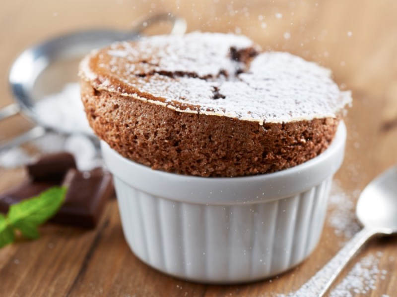 Ein schokoladensoufflé in einer kleinen Form, das mit Puderzucker bestreut wurde. Daneben ein Löffel, einige Stücke Schokolade, ein Sieb und frische Minze.