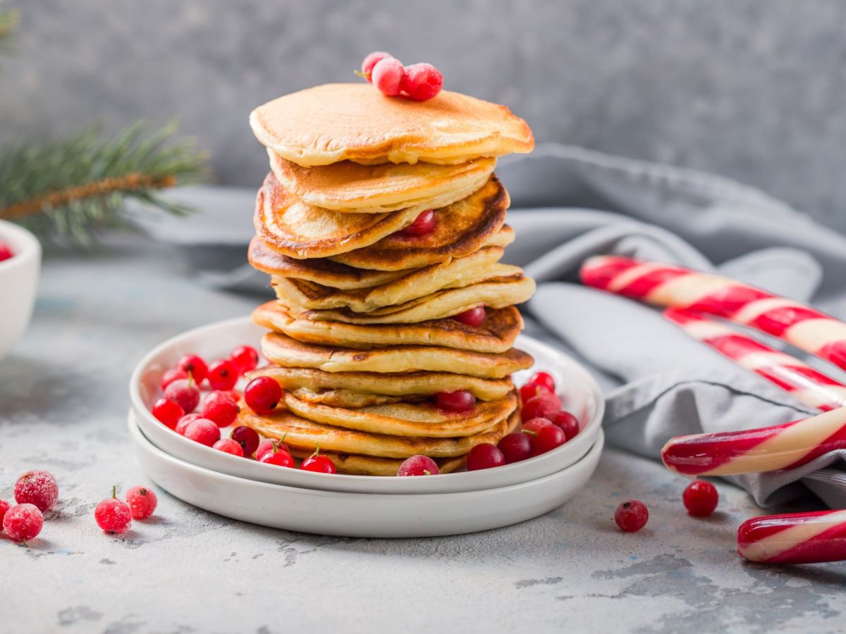 Ein Stapel winterliche Pancakes liegt auf einem Teller. Bestreut sind sie mit Puderzucker und Preiselbeeren. Als Deko liegen Preiselbeeren und Zuckerstangen neben dem Teller. Im Hintergrund liegt eine graue Tischdecke.