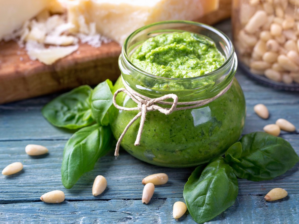 Ein Glas Basilikumpesto verziert mit einer Schleife, darum herum BasilikumblÃ¤tter und Pinienkerne.