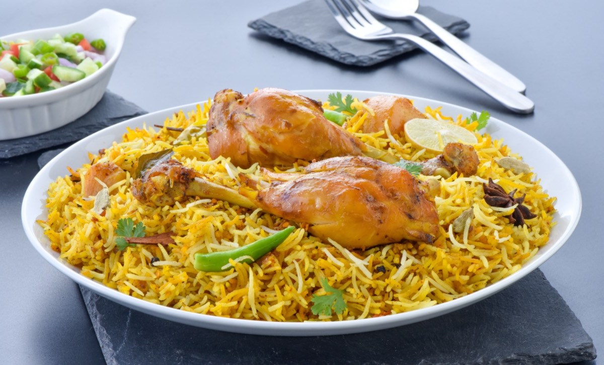 Ein tiefer Teller mit Chicken Biryani. Im hintergrund sind Besteck und eine Schale mit Salat.