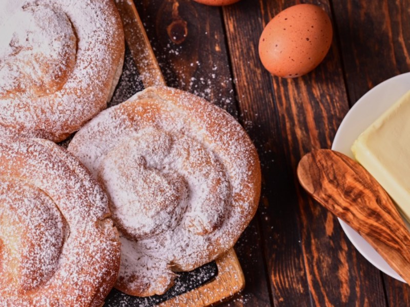 Drei Ensaimadas auf einem Holzbrett. Daneben liegen ein Ei und ein Teller mit einem Stück Butter und einem Holzlöffel darauf.