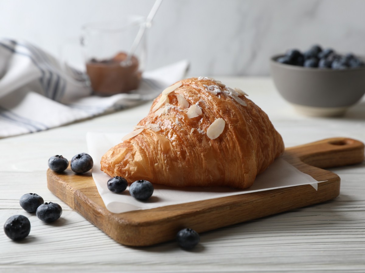 Ein gefÃ¼lltes Mandelcroissant mit Mandeln und Puderzucker verziert auf einem Brett, rundherum Blaubeeren.