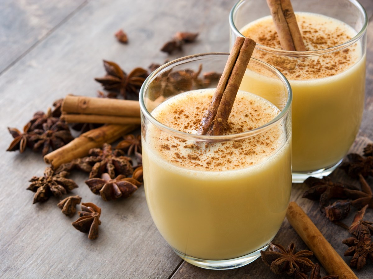 2 Gläser Heiße Oma, ein Getränk mit Milch und Eierlikör, garniert mit einer Zimtstange und geriebenem Zimt, drumherum winterliche Gewürze.