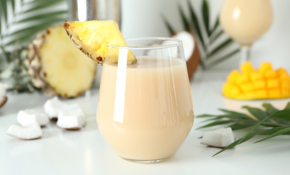 Ein Glas heiße Piña Colada mit einem Stück frischer Ananas als Deko vor Palmenblättern, frischer Ananas und Mango im Hintergrund.