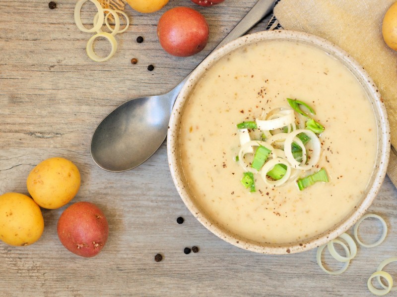 Eine Schüssel Kartoffel-Lauch-Suppe mit Lauch und Pfeffer garniert, daneben ein Löffel und Kartoffeln, Draufsicht.