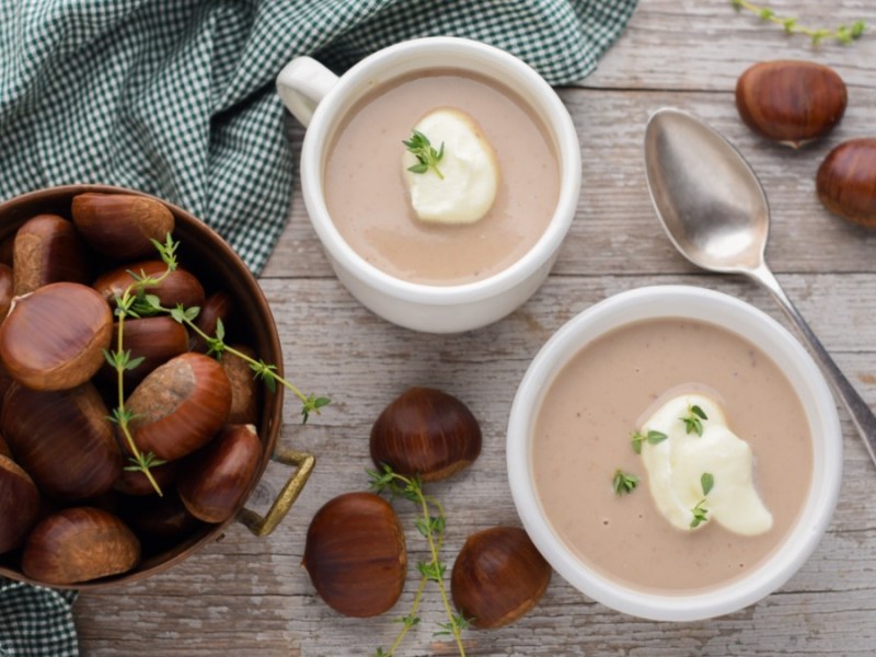 Zwei kleine Suppenterrinen mit Maronen-Cremesuppe, die mit Thymian und einem Klecks Soja-Kochcreme garniert sind. daneben steht eine Schale mit Marone und es liegen einige drumherum.