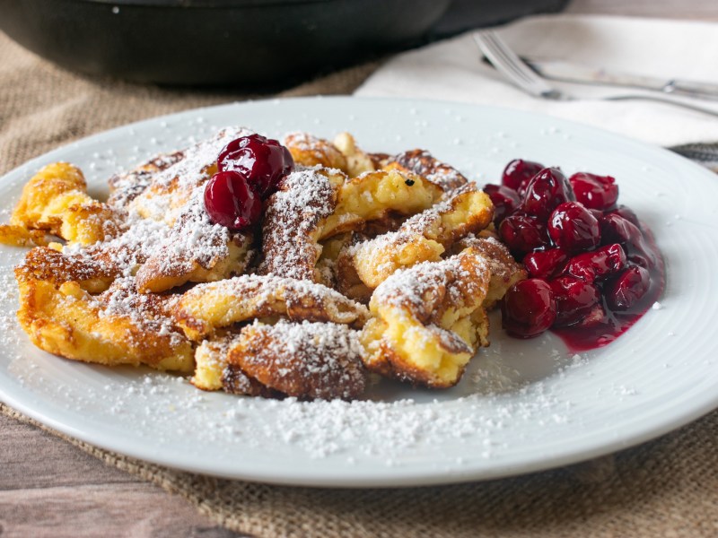 Österreichischer Kaiserschmarrn auf einem weißen Teller, serviert mit Kirschen und Puderzucker