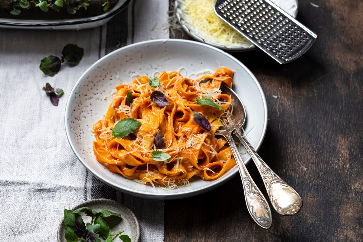 Ein Teller mit Paprika-Pasta mit Käse und Basilikum garniert, darauf Besteck, daneben eine Schale mit Käse und eine Reibe.