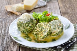 2 Spinatknödel, einer halbiert, auf einem Teller mit Salat, im Hintergrund ein Stück Parmesan, neben dem Teller Besteck.