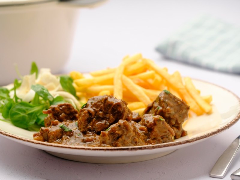 Ein Teller mit Stoofvlees, traditionelles belgisches Bier-Gulasch, mit Pommes und Salat. Im Hintergrund steht ein Bräter.