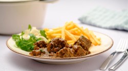 Ein Teller mit Stoofvlees, traditionelles belgisches Bier-Gulasch, mit Pommes und Salat. Im Hintergrund steht ein Bräter.