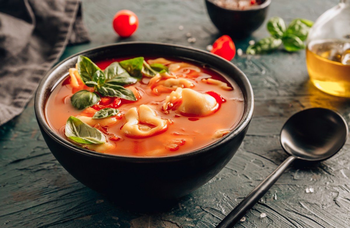 Eine schwarze Schüssel Tortellini-Suppe mit roter Tomatensuppe und Basilikumblättern, daneben ein Löffel, eine Flasche mit Öl und wenige weitere Zutaten.