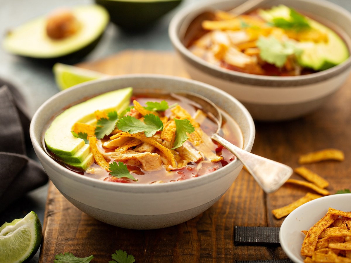 Zwei SchÃ¼sseln Tortilla-Suppe mit Avocado und Tortilla-Streifen, drumherum verschiedene Zutaten wie eine halbierte Avocado.