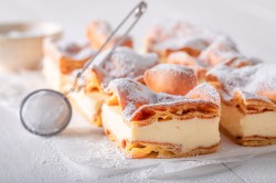 Blätterteigkuchen mit Vanillepudding auf einer weißen Marmorplatte. Darauf liegt ein Sieb, mit dem man den Kuchen mit Puderzucker bestreuen kann.