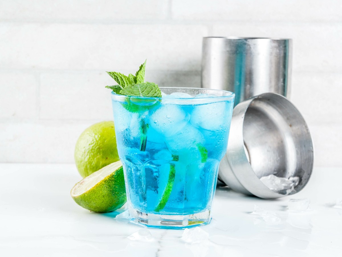 Ein Glas Blue Lagoon mit EiswÃ¼rfeln, Limettenscheiben und Minze garniert, dahinter ein Cocktailshaker und halbierte Limetten.