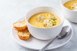 Eine Schüssel Blumenkohl-Kartoffel-Suppe mit geröstetem Blumenkohl garniert auf einem Teller, auf dem auch ein Löffel und zwei Scheiben getoastetes Weißbrot liegen.