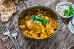 Chicken Korma in einer Metallschale mit Koriander garniert. Daneben liegen Naan-Brot, zwei Gabeln und eine Schale gemahlene Mandeln.