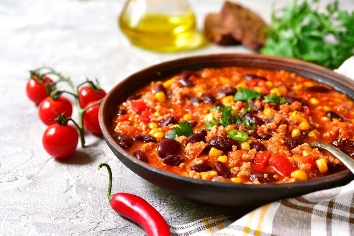 Ein tiefer Teller Chili con Carne mit Schokolade, darin ein Löffel, daneben Tomaten an der Rispe und eine Chilischote.