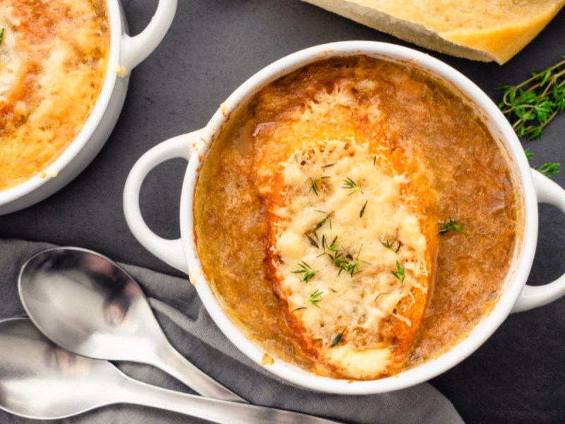 Eine Terrine mit französischer Zwiebelsuppe. Es befinden sich eine weitere Schüssel und zwei Suppenlöffel daneben.