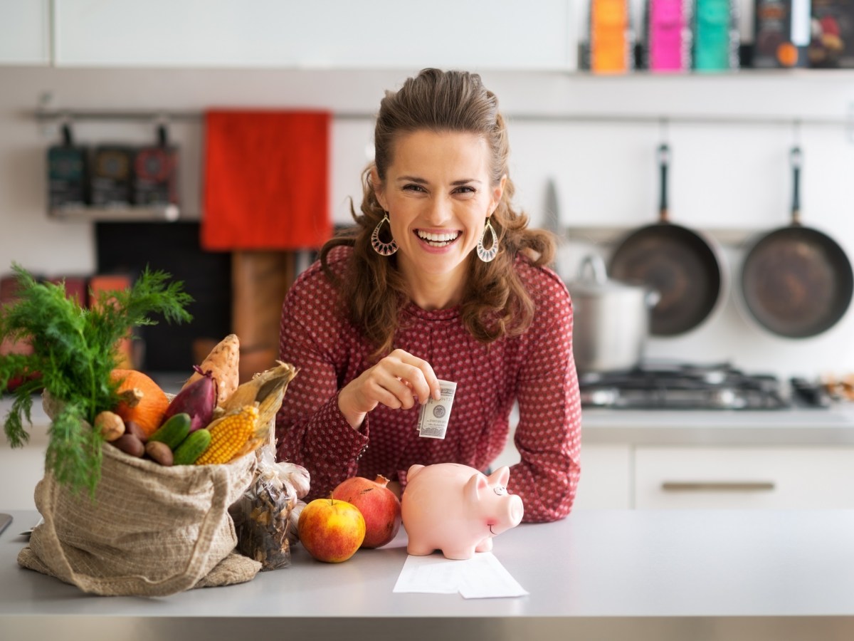 Geld sparen beim Kochen: eine lachende, lockige Frau steht in der KÃ¼che, steckt einen Geldschein in ein Sparschwein, neben ihr liegen EinkÃ¤ufe.