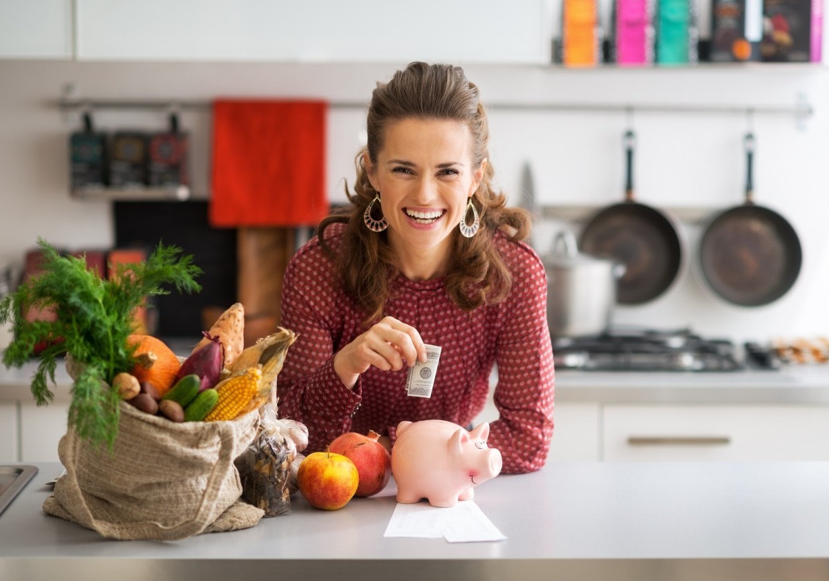 Geld sparen beim Kochen: eine lachende, lockige Frau steht in der Küche, steckt einen Geldschein in ein Sparschwein, neben ihr liegen Einkäufe.