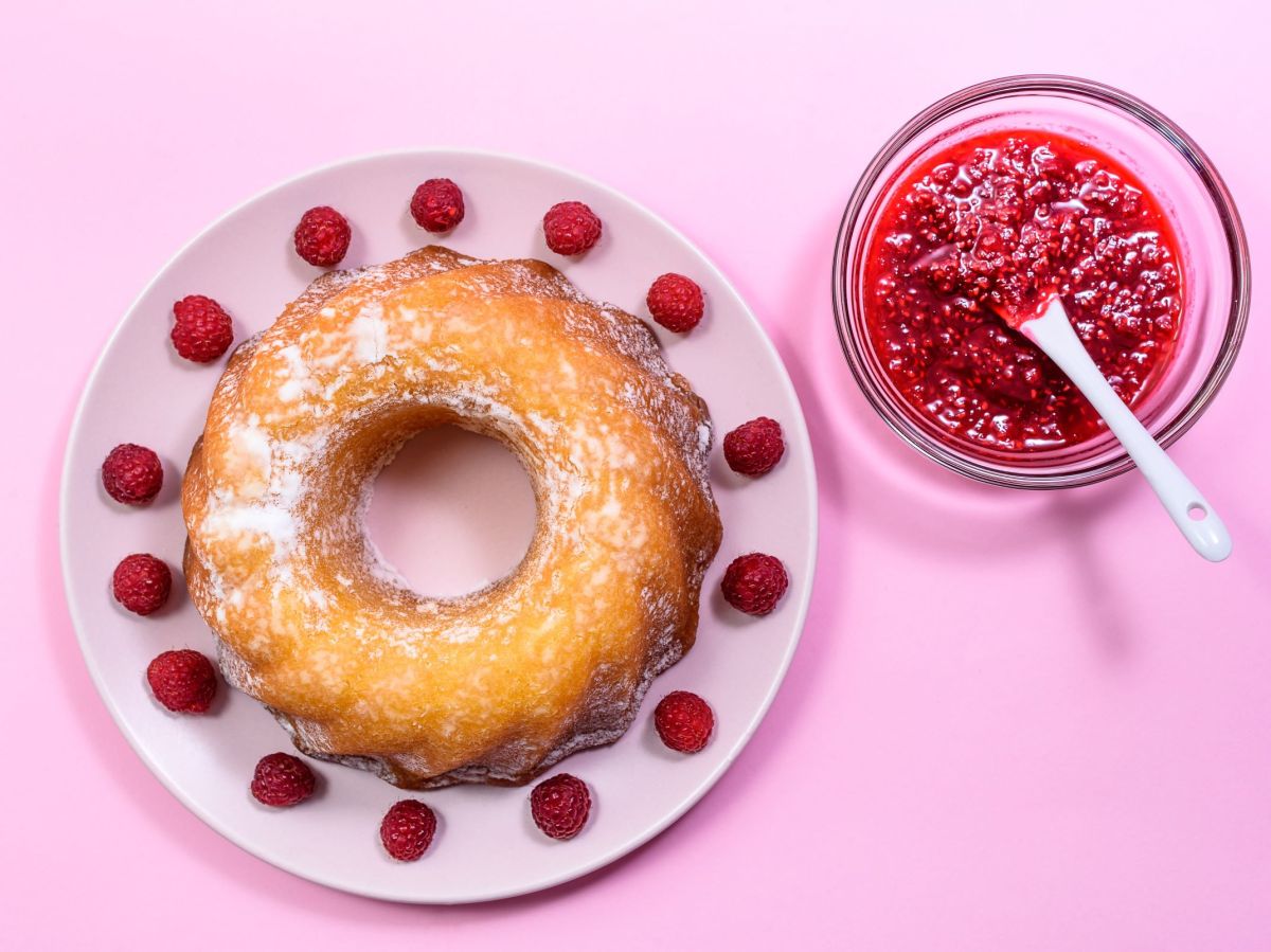 Himbeer-Zitronen-Gugelhupf, angerichtet auf einem Teller und verziert mit frischen Himbeeren. Ein Schälchen mit Himbeerkompott steht daneben.