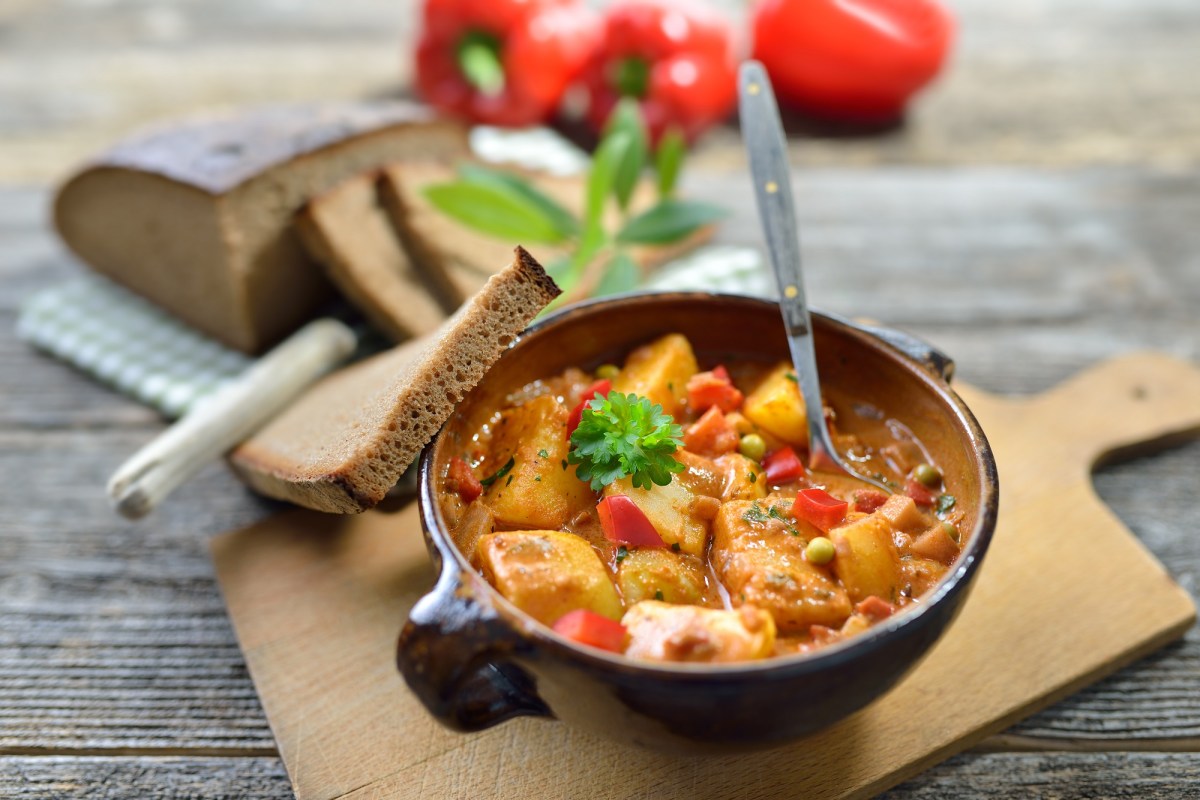 Getöpferte Schale mit Kartoffel-Gulasch auf einem Brett und drei Scheiben Brot daneben.