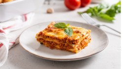 Ein Stück klassische Lasagne auf einem weißen Teller. Im Hintergrund sind frische Tomaten, Kräuter und die restliche Lasagne zu sehen.