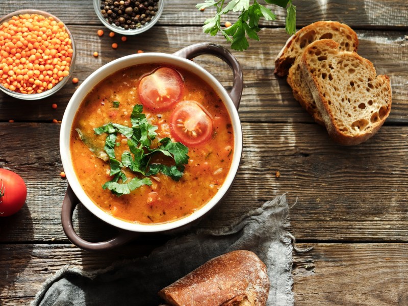 Eine Schüssel mit Henkeln, darin Linsen-Tomaten-Suppe, garniert mit halbierten Tomaten und Koriander, daneben frische Tomaten, Brotscheiben, eine Schale Linsen, Draufsicht.