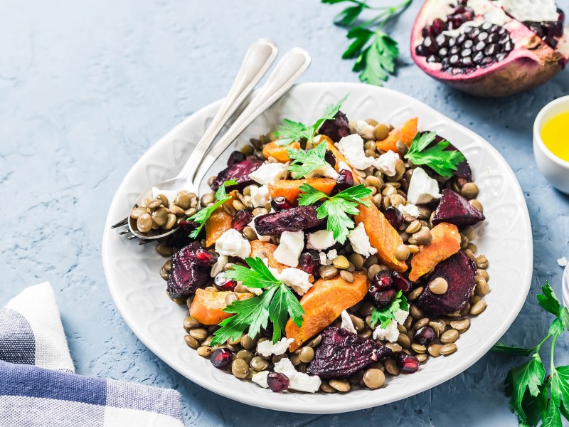 Linsensalat mit Wintergemüse auf einem weißen Teller. Daneben Petersilie und eine Schale Olivenöl.