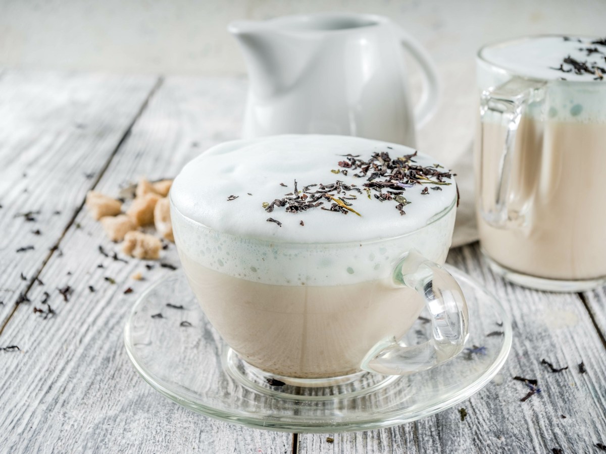 Durchsichtige Tasse London Fog Latte, Milchschaum und Lavendelblüten auf einem grauen Untergrund.