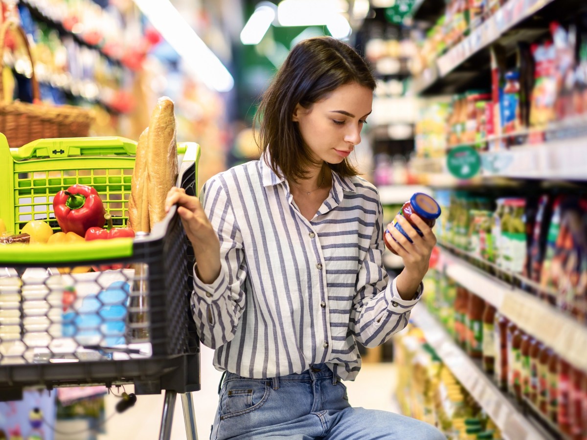 Eine dunkelhaarige junge Frau prÃ¼ft im Supermarkt das Mindesthaltbarkeitsdatum eines Lebensmittels im Glas und hockt dabei neben ihrem Einkaufswagen.