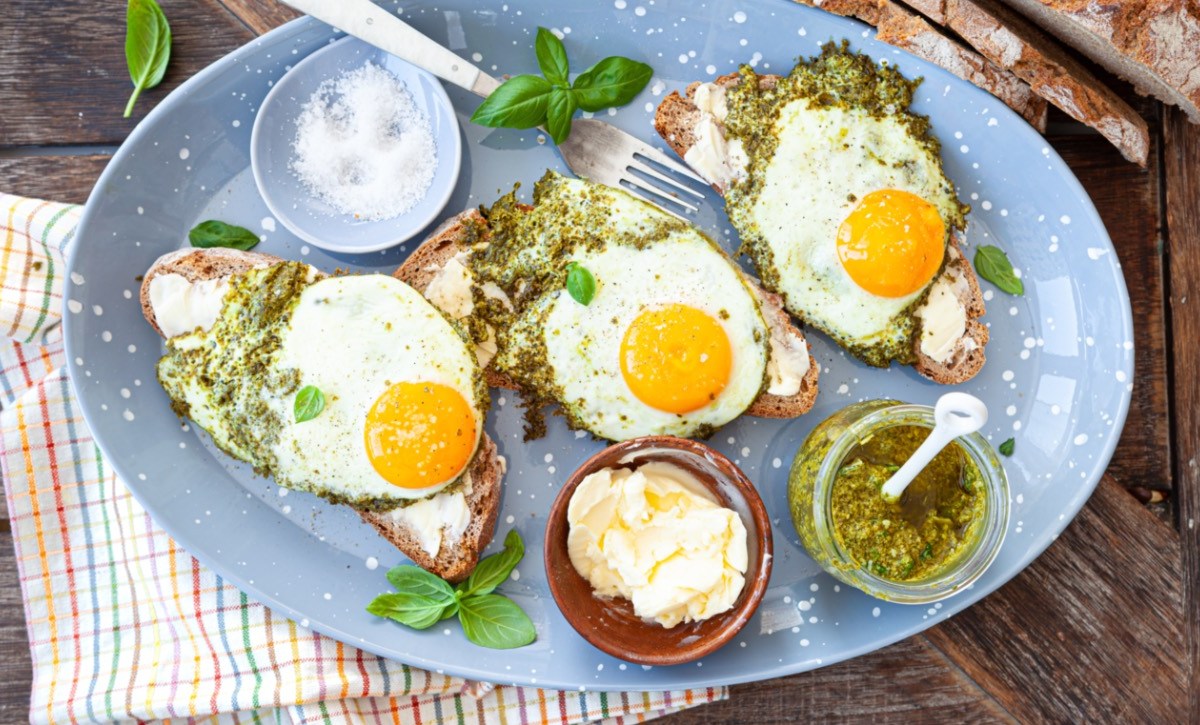 Drei Pesto-Eier auf Brotscheiben. Drumherum stehen Schalen mit Pesto, Butter und Salz.